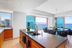 A modern kitchen with a large island, a dining area with round tables, and large windows offering ocean views. - Puerto Vallarta 3BR Ocean View Condo w/ Pool & Gym (Puerto Vallarta)