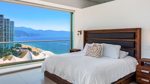 Ein Schlafzimmer mit großem Fenster, einem Holz-Kopfteil und Blick auf Strand und Berge.