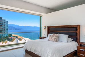 A bedroom with a large window, a wooden headboard, and a view of a beach and mountains. - Puerto Vallarta 3BR Ocean View Condo w/ Pool & Gym (Puerto Vallarta)