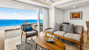 Een moderne woonkamer met een bank, stoelen, een salontafel en een plafondventilator. Er is een groot raam met uitzicht op de oceaan.