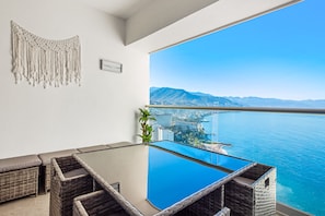 Ein Balkon mit Glastisch und Korbmöbeln, mit Blick auf eine Küstenstadt und das Meer.