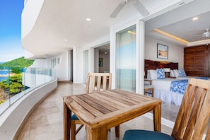 Ein Balkon mit Holztisch und -stühlen, ein Bett mit blau-weißer Bettwäsche und Blick auf eine grüne Landschaft.
