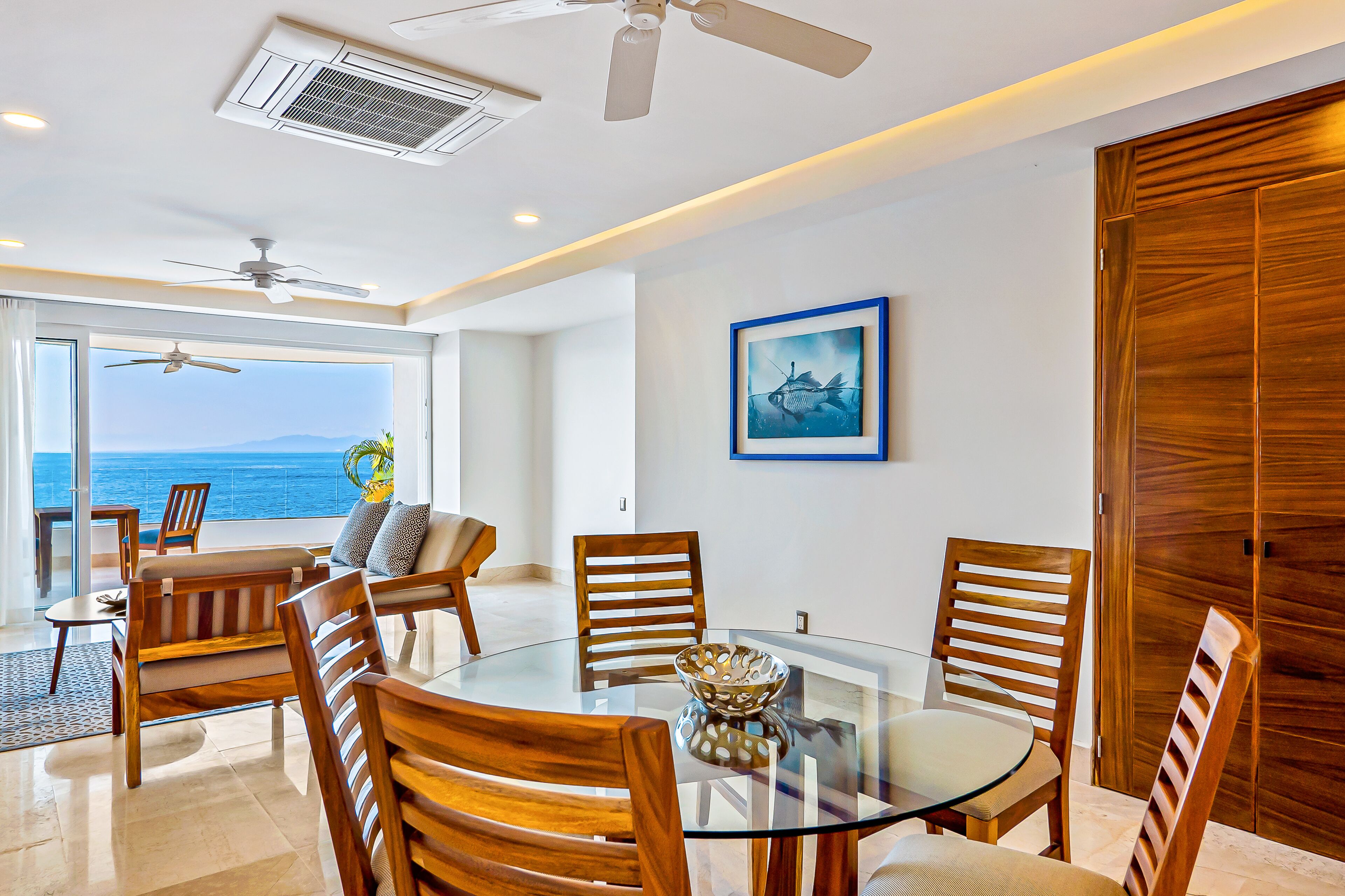 A modern dining area with a glass table, wooden chairs, and a view of the ocean.