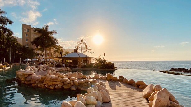 A wooden deck leading to a pool area with large rocks, palm trees, and a building in the background. - Beachfront dream retreat with pool, tennis & gym access - washer/dryer, AC (Puerto Vallarta)