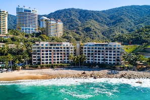 Un hotel frente a la playa con varios pisos, una playa en primer plano y montañas al fondo.