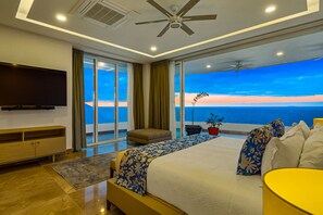 Een moderne slaapkamer met een groot bed, een televisie, een plafondventilator en een balkon met uitzicht op de oceaan.