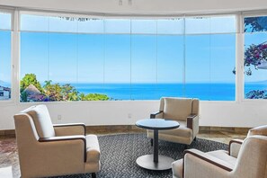 A room with a large window offering a view of the sea, two armchairs, a small round table, and a patterned carpet. - Modern hillside suite with balcony, city view, pools, gym & full kitchen (Puerto Vallarta)