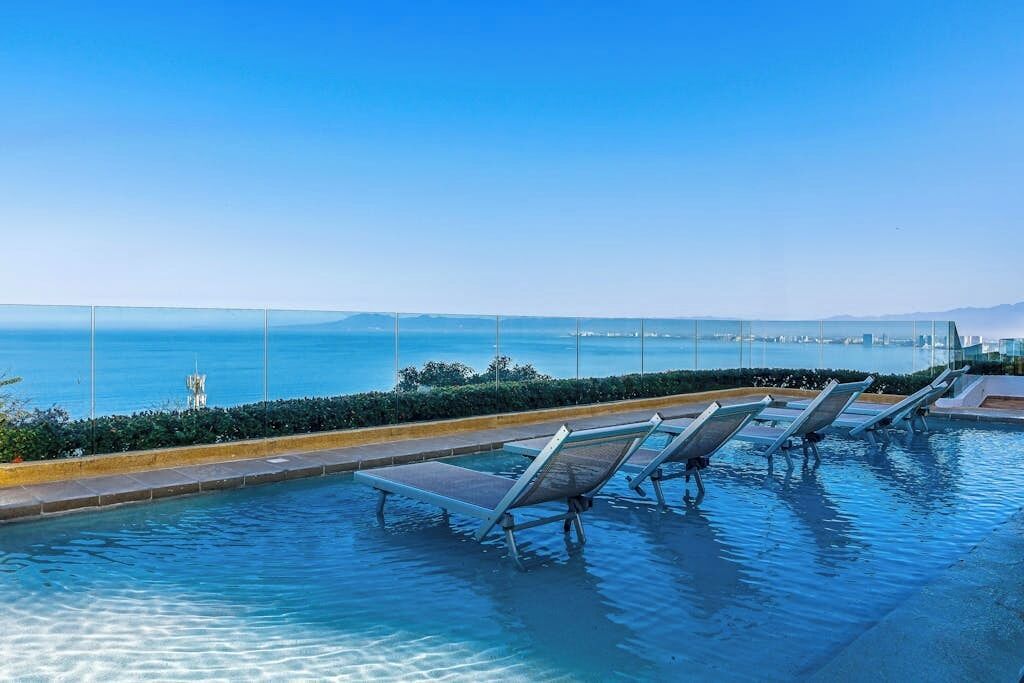 Una piscina con tumbonas, vista despejada al mar y un paisaje urbano a lo lejos.