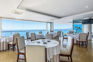 Een eetruimte met ronde tafels, stoelen en een groot raam met uitzicht op zee.