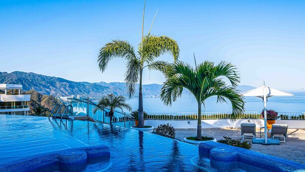Une piscine à l’eau cristalline, bordée de palmiers, et offrant une vue sur la mer et les montagnes.