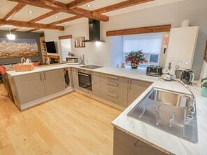 A modern kitchen with wooden beams, a large island, a built-in oven, and a window with a view. - May Cottage, Snainton (Scarborough)