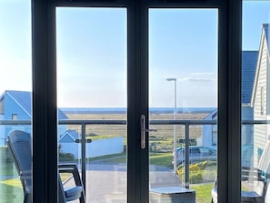 A view through a glass door with a metal handle, leading to a balcony with a clear sky and distant buildings.