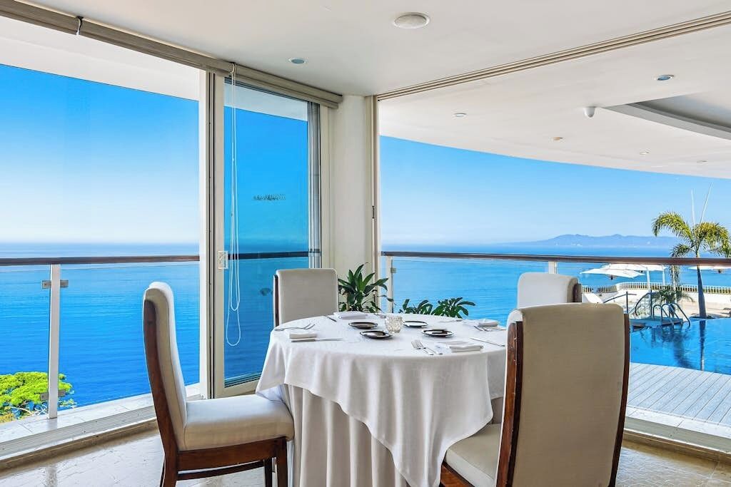 Comedor con mesa lista para cuatro personas, con vista a una piscina y al mar.