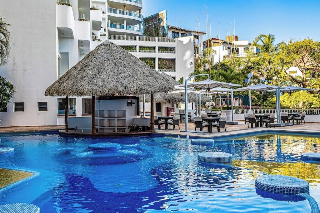 Una piscina con techo de paja, rodeada de sillones de descanso y sombrillas, con edificios modernos al fondo.