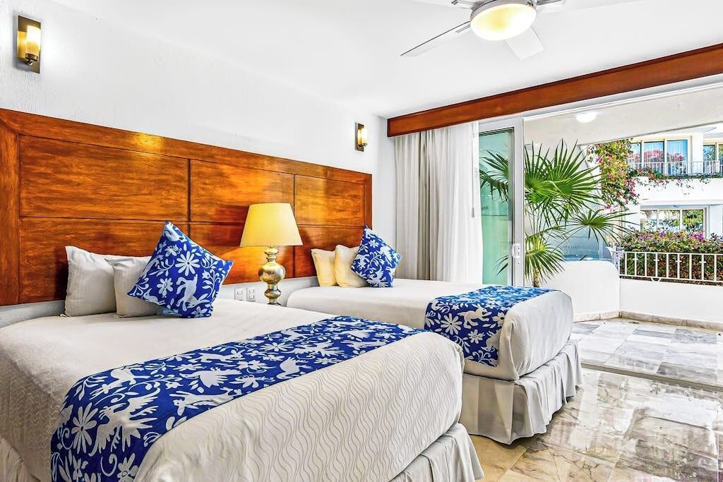 Ein Hotelzimmer mit zwei Betten, einem Holz-Kopfteil, einem Deckenventilator und einem Balkon mit Pflanzen.