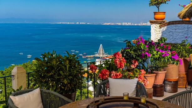 A seaside terrace with a table, chairs, potted plants, and a view of the sea and distant cityscape. - Paraiso del Mar 1BR/1BA Ocean View in Puerto Vallarta (Puerto Vallarta)