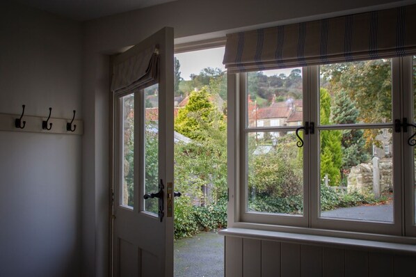 Une pièce avec une porte donnant sur un jardin, une fenêtre offrant une vue sur la verdure et les bâtiments, et un porte-manteau avec des crochets.