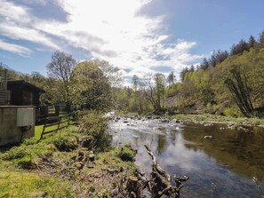 A river flowing through a forested area with a wooden cabin on the left. - Elm Lodge, Lake District, 2bed (Keswick)
