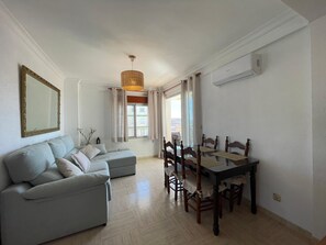 A living room with a sofa, dining table, chairs, a hanging light fixture, and a window with curtains. - Terrasol Madrid 9 in Torre del Mar (Velez-Malaga)