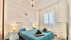 Een slaapkamer met een bed, nachtkastje, lamp, raam met gordijnen en een kledingkast.