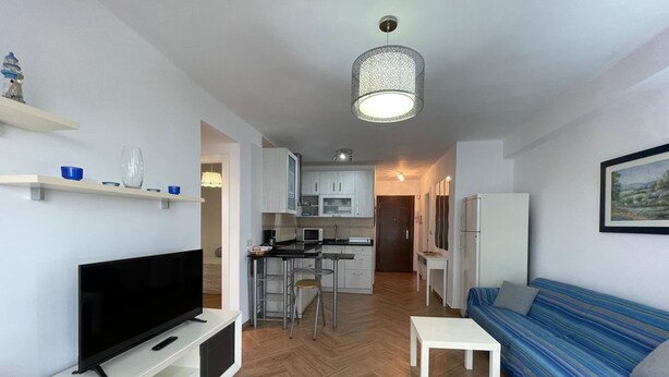 A modern living room with a flat-screen TV, a sofa, a small table, and a kitchen in the background. - Terrasol SAN Francisco Larios (Velez-Malaga)