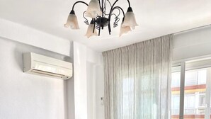 Ein modernes Zimmer mit Esstisch, vier Stühlen, einem Glastisch, einer Couch, einer Kronleuchter und Blick ins Freie durch ein großes Fenster.