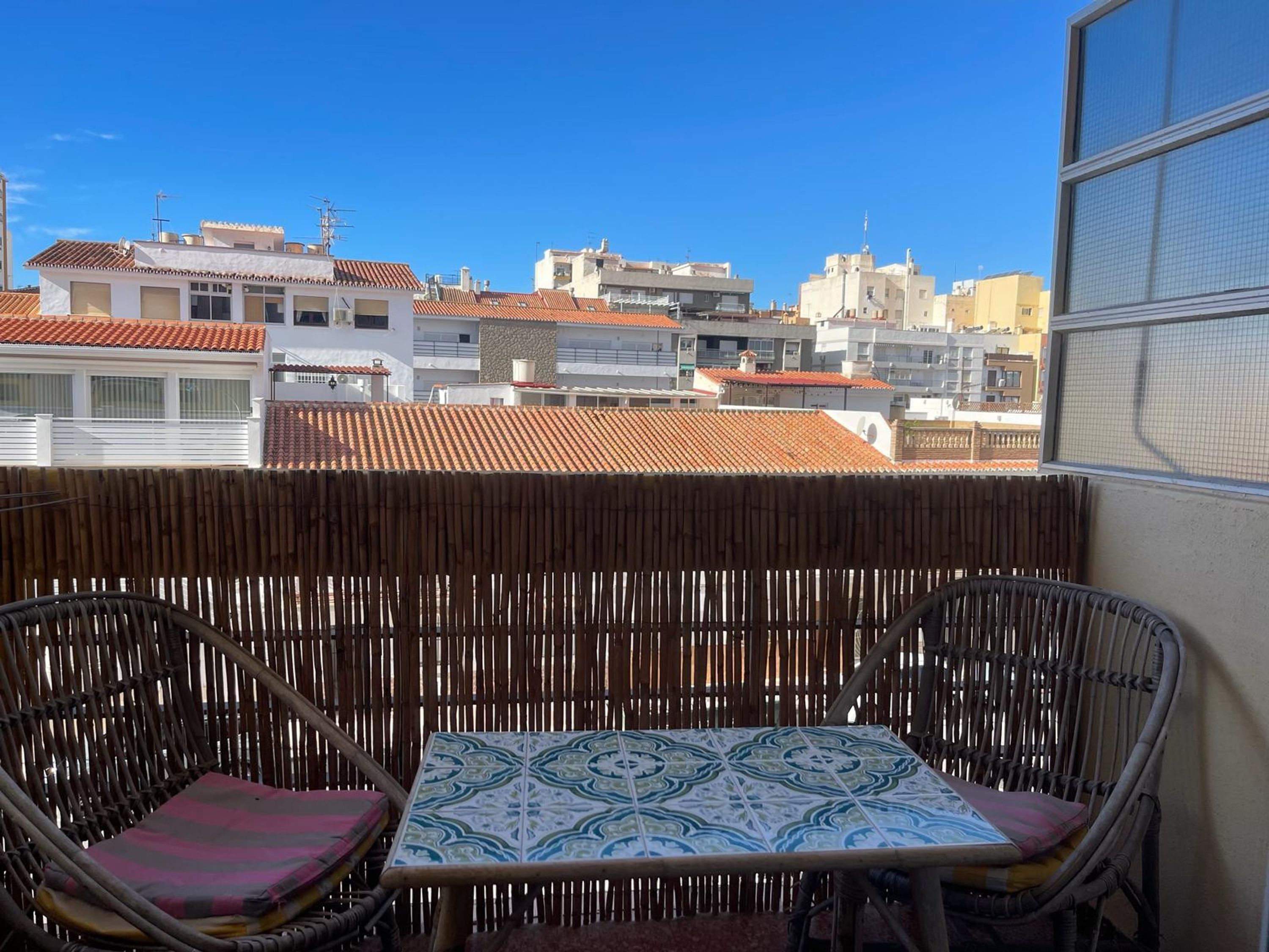 Ein Balkon mit Korbmöbeln und einer gemusterten Tischdecke, mit Blick auf eine Stadtlandschaft mit Terrakottadächern.