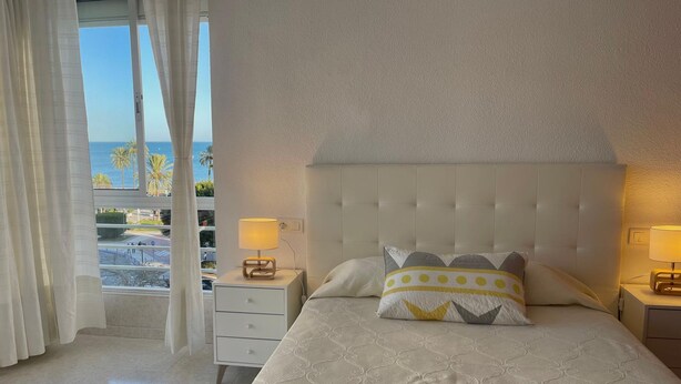 A bedroom with a bed, two bedside tables, a nightstand, a window with curtains, and a view of the sea. - Terrasol California 4 (Velez-Malaga)