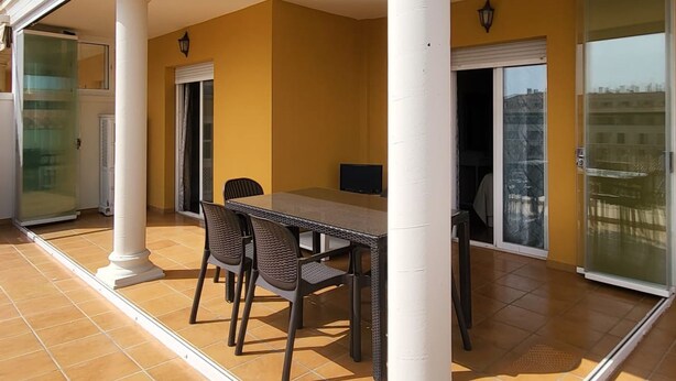Un patio techado al aire libre con una mesa y sillas, un vidrio corredizo y un balcón con barandillas rojas.