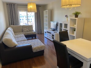 Ein Wohnzimmer mit Ecksofa, Esstisch, Fernseher und Bücherregal.