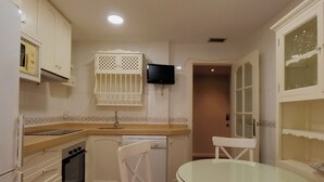 Una cocina compacta con armarios blancos, microondas integrado, lavavajillas y televisor montado en la pared.