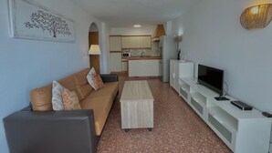 Ein Wohnzimmer mit Sofa, Couchtisch, Fernseher und einer Küche im Hintergrund.