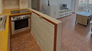 Eine kompakte Küche mit Herd, Backofen und Kühlschrank. Es gibt einen Essbereich mit Tisch und Stühlen sowie einen Blick ins Freie durch ein Fenster.