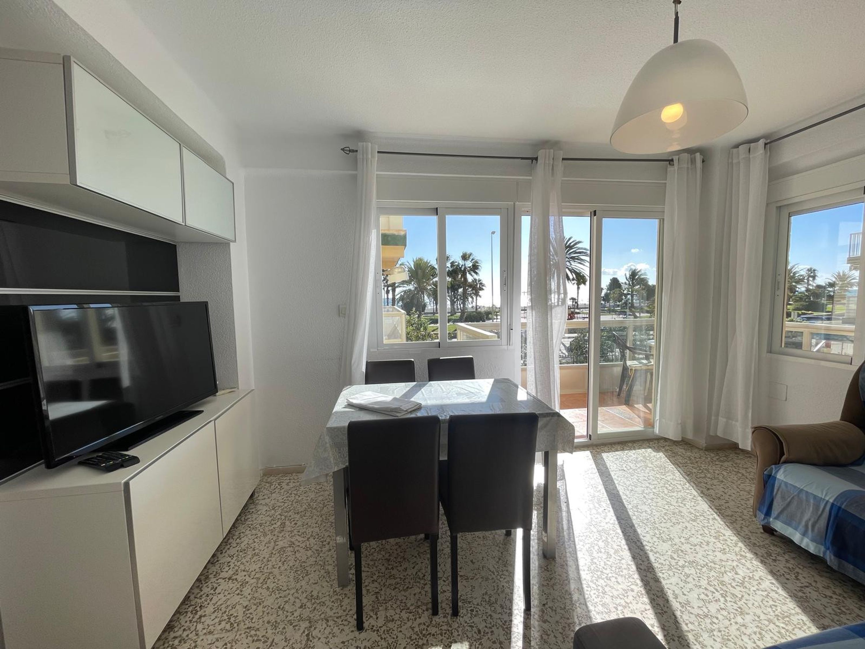 A modern kitchen with a flat-screen TV, a dining table with chairs, and a balcony with a view of palm trees.