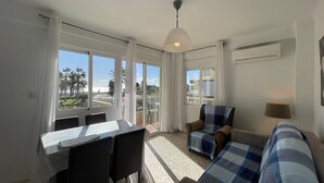 A room with a dining table, chairs, a sofa, and a view of palm trees and buildings. - Terrasol SAN Gerardo 2-1-3 Frontal AL MAR (Velez-Malaga)