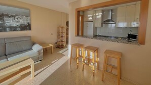 A living room with a sofa, a small table, and three wooden stools. A kitchen with white cabinets and a granite countertop is visible through an open doorway. - Terrasol Puerto Blanco 4d-10 (Velez-Malaga)