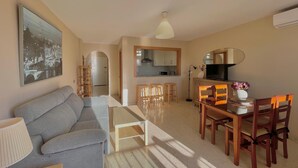 A living room with a sofa, a dining table with chairs, a TV, and a kitchen in the background. - Terrasol Puerto Blanco 4d-10 (Velez-Malaga)