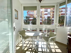A sunlit room with a glass door, two chairs, a small table, and a view of multi-story buildings. - Terrasol Plazamar 3-6 (Velez-Malaga)