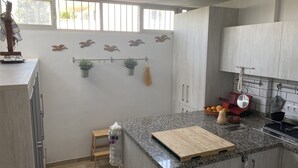 A kitchen with granite countertops, wooden cabinets, a small wooden stool, a cutting board, and a window with blinds. - Terrasol Trai AS A1- 1 Linea Playa (Velez-Malaga)