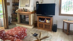 A living room with a fireplace, a television, a bookshelf, and a window with a view of greenery. - Terrasol Trai AS A1- 1 Linea Playa (Velez-Malaga)