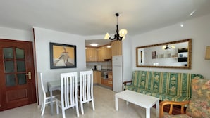 A living room with a sofa, a dining table with chairs, a kitchen in the background, a door, and a mirror on the wall. - Terrasol Caracola 3 in Torre del Mar (Velez-Malaga)