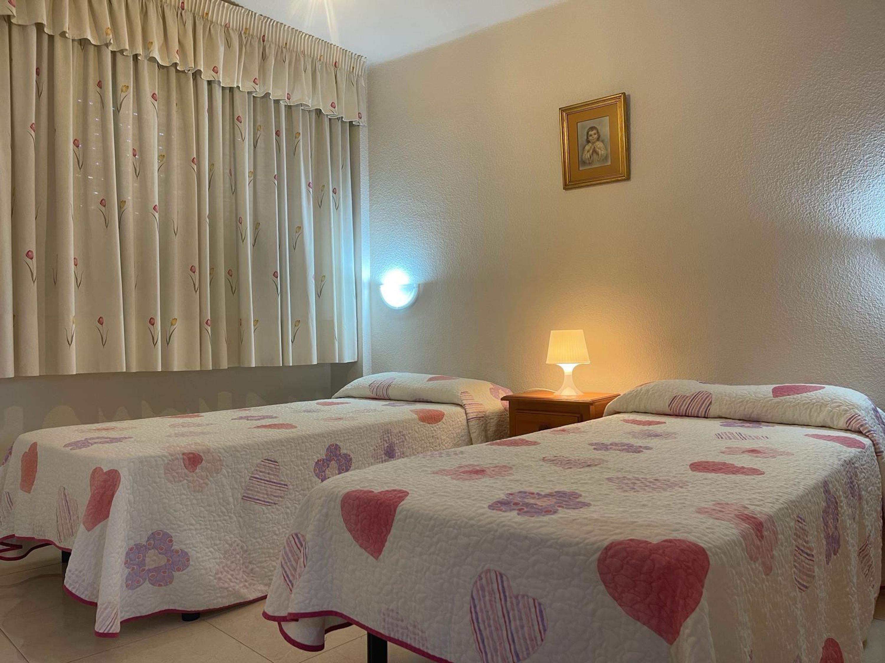 A hotel room with two beds, a nightstand with a lamp, a wall-mounted picture, and a window with curtains.