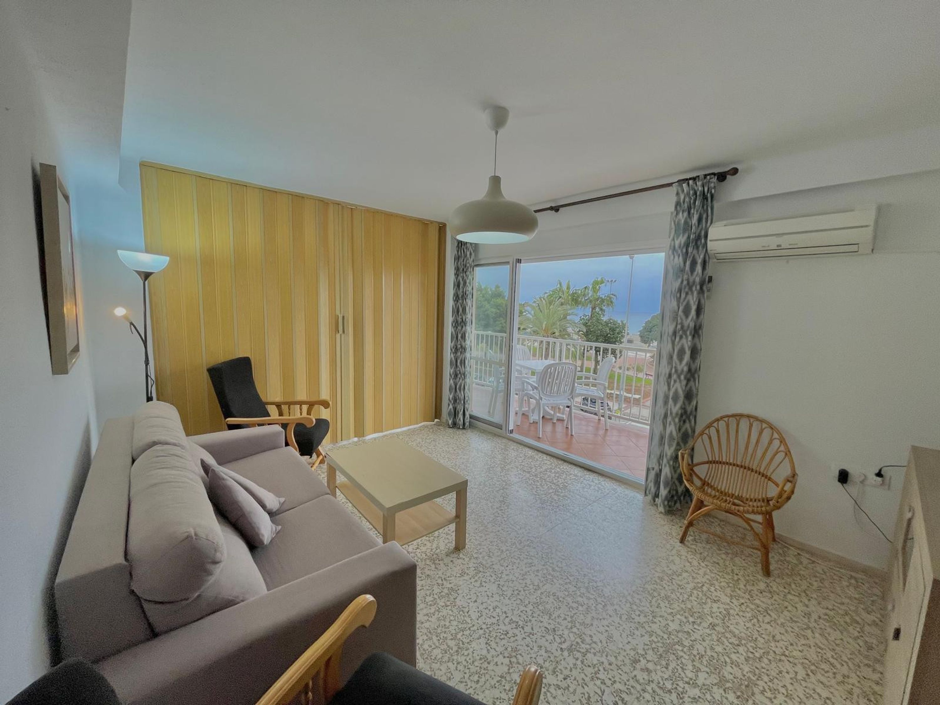 A living room with a sofa, a chair, a table, and a balcony with a view.