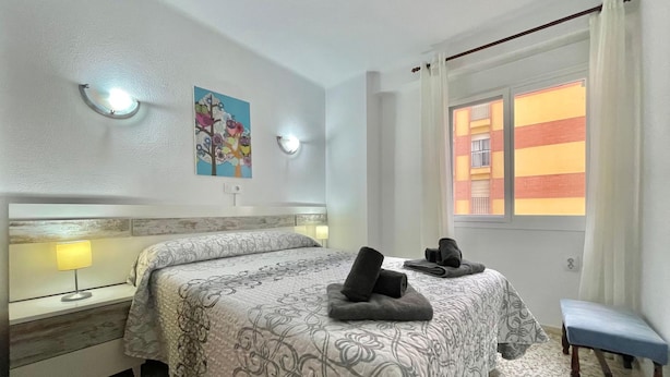 A hotel room with a bed, bedside lamps, a bench, and a window with a view of buildings. - Terrasol SAN Gerardo 2-2-2 Frontal AL MAR (Velez-Malaga)