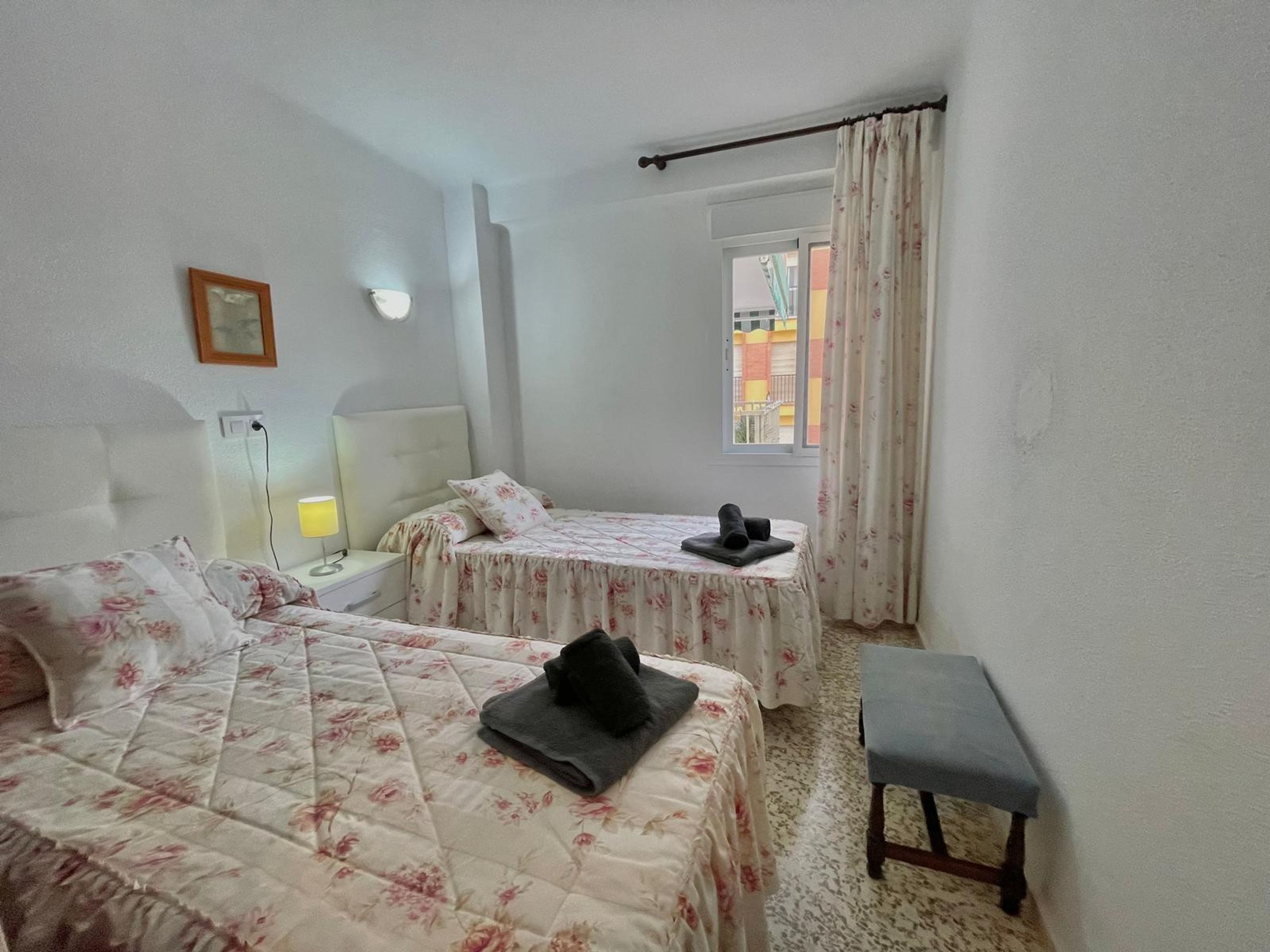 A hotel room with two beds, a bedside table, a bench, a window with curtains, and a wall-mounted light.