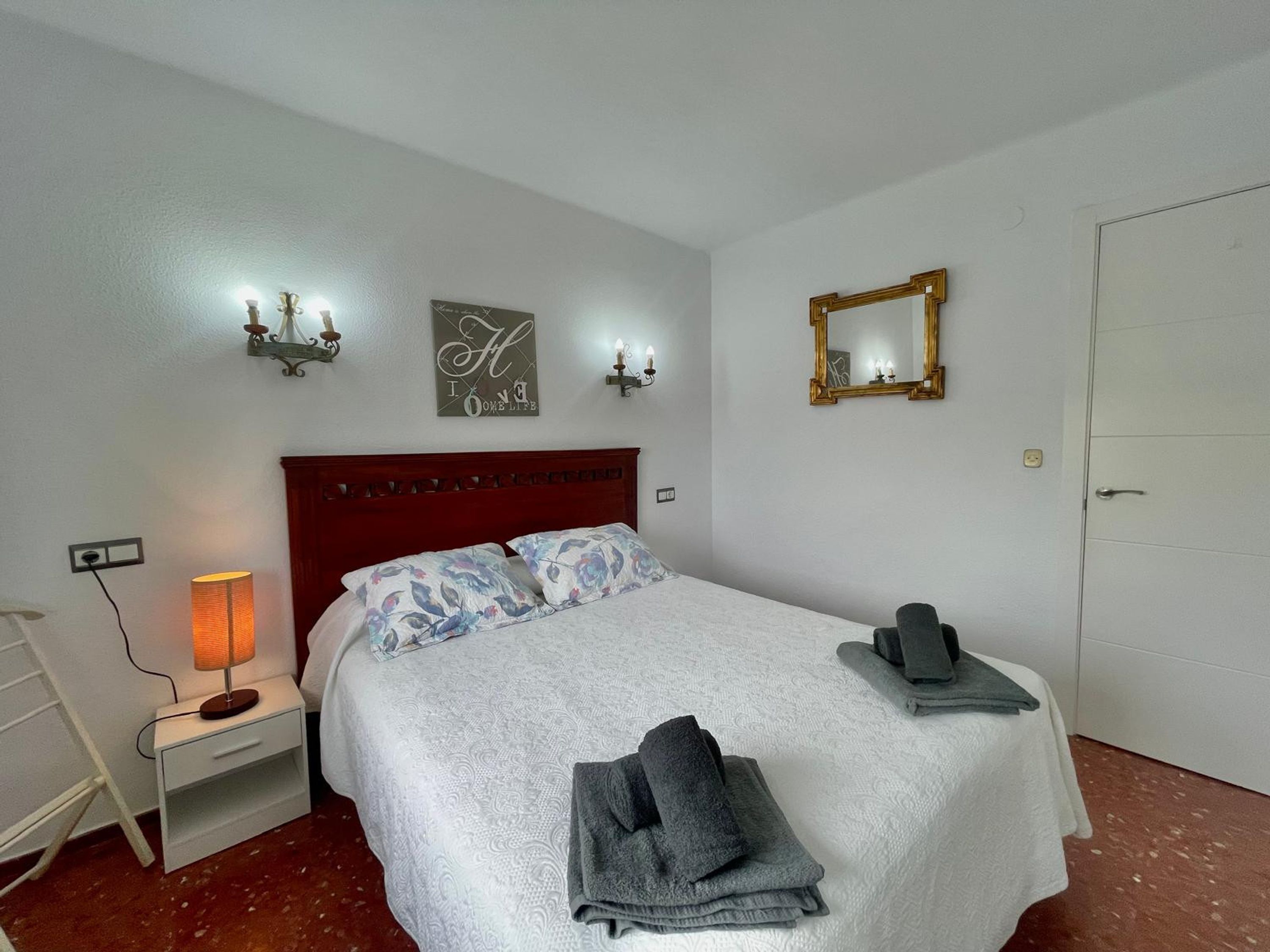 A neatly made bed with a white bedspread and floral pillows, a bedside lamp, a folded towel, a mirror, and a wall with decorative wall art.