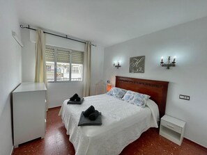 Une chambre à coucher comprenant un lit, une table de chevet, une commode, une fenêtre avec des stores et un luminaire fixé au mur.