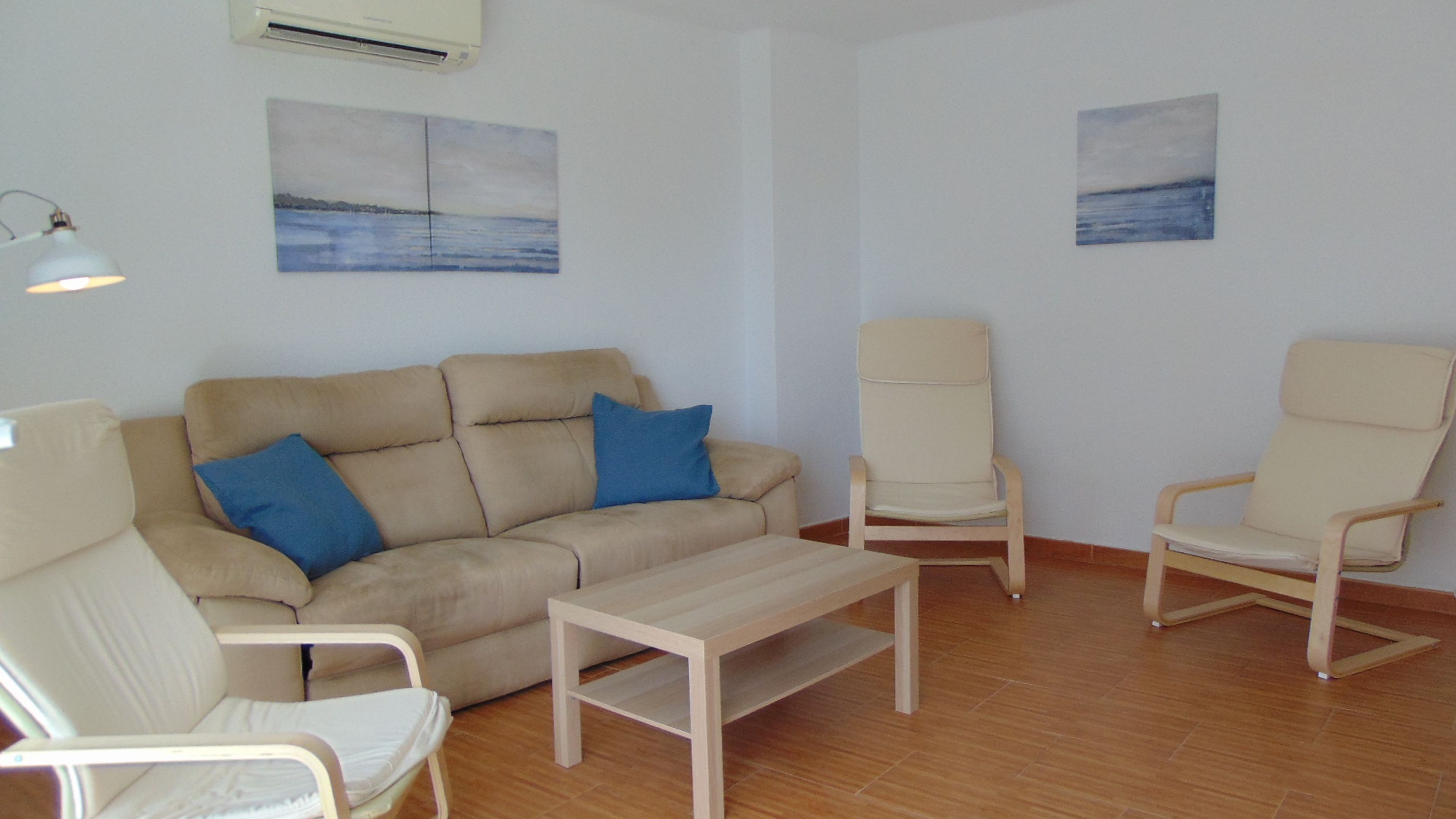 Una sala de estar con un sofá beige, dos sillones, una mesita y dos cuadros colgados en la pared.