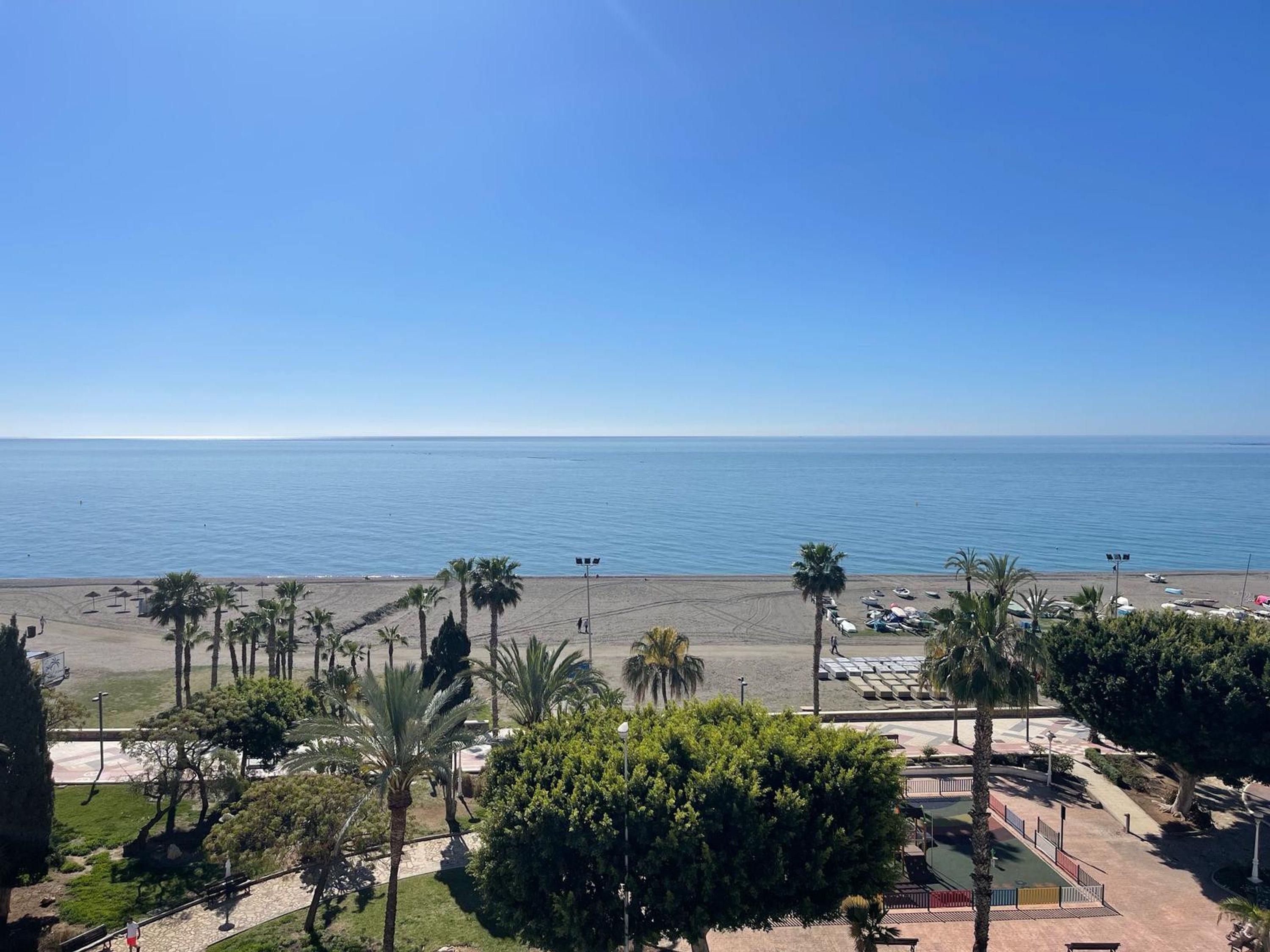 Blick auf einen Strand mit Palmen, einem gepflasterten Weg und einem Parkplatz.