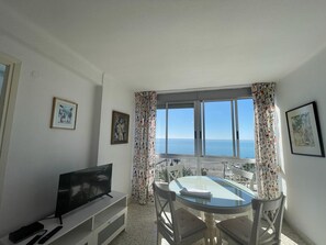 A room with a TV, a table with chairs, a window with a view of the sea, and framed pictures on the walls. - Terrasol Frontal AL MAR Antillas 8 (Velez-Malaga)